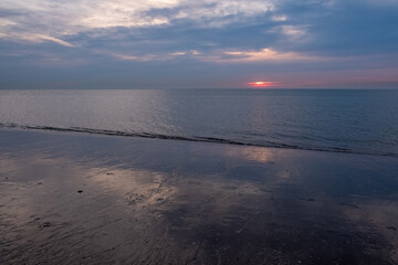 The image conveys a serene sunset at low tide, where the receding water leaves wet sand behind, mirroring the dwindling light. The sun, a fiery orb, dips towards the horizon, its warm glow contrasting