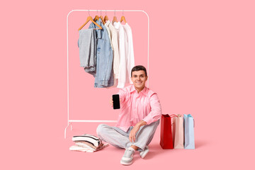 Handsome young happy man holding mobile phone and rack with stylish clothes on pink background