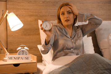 Mature woman with alarm clock cannot sleep in bedroom at night. Menopause concept