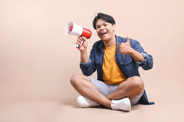 Young Asian Man Sitting on The Floor Shouting Through a Megaphone While Showing Thumb Up Isolated...