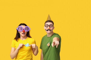 Young friends in funny disguise with paper fishes pointing at viewer on yellow background. April...