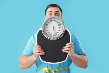Shocked young man with tape measure around his waist and scales on blue background. Weight loss...