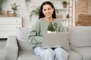 Young happy woman sitting on sofa with houseplant and laptop in living room