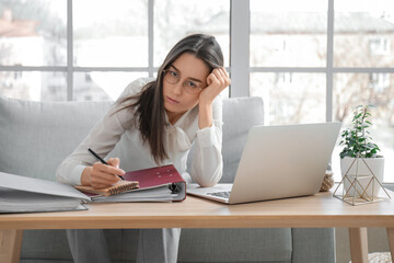 Tired young businesswoman sitting at table with laptop and office folders in living room