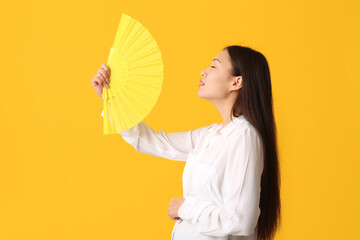 Young woman with hand fan suffering from heat on yellow background