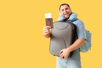 Young happy man with suitcase, headphones, passport and travel pillow on yellow background
