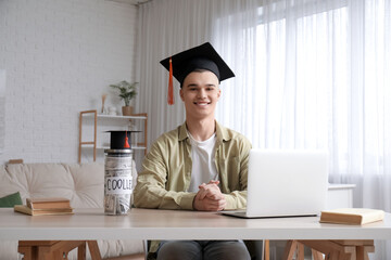 Male student in graduation hat with laptop at table with jar bank in room. Concept of savings for...