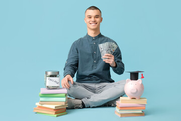Male student sitting with piggy bank in graduation hat and books on blue background. Concept of...