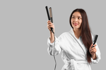 Beautiful young happy Asian woman in bathrobe with brush and straightening iron on grey background