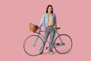 Beautiful young Asian woman with bicycle and wicker basket on pink background - obrazy, fototapety, plakaty