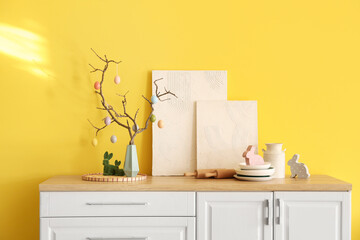 Vase with tree branches, Easter eggs, paintings and kitchen utensils on counters near yellow wall