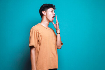 Tired Asian guy standing and yawning isolated on teal background