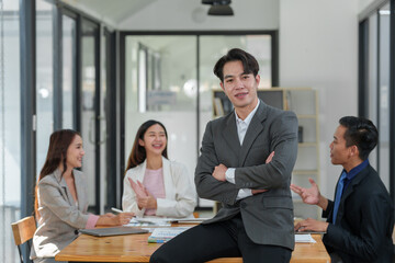 Strong young Asian businessman A muscular man smiles at the camera while leaning against his desk, arms crossed happily in the conference room of a modern workplace.