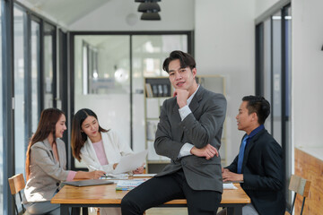 Fototapeta na wymiar Strong young Asian businessman A muscular man smiles at the camera while leaning against his desk, arms crossed happily in the conference room of a modern workplace.