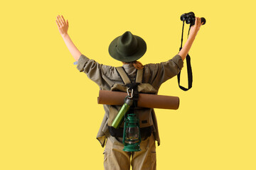 Beautiful young female tourist with binoculars and backpack on yellow background, back view