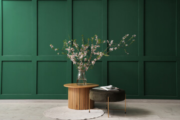 Pouf and vase with blooming branches on coffee table near green wall