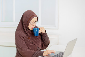 Elderly Asian woman drinking coffee or tea and working from laptop at her home