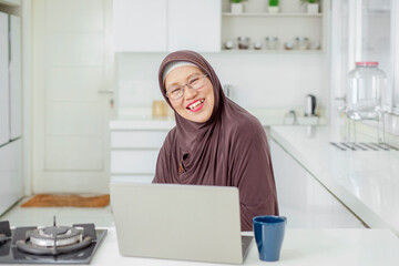 Happy Elderly Asian Muslim woman drinking coffee or tea and working from laptop at her home