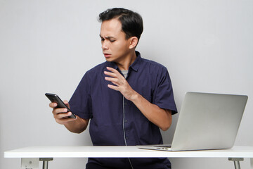 angry asian man holding mobile phone while sitting in front of laptop computer. on isolated...