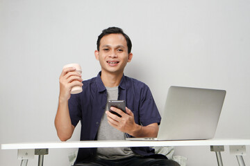 joyfull happy asian man holding mobile phone while sitting in front of laptop computer. on isolated background