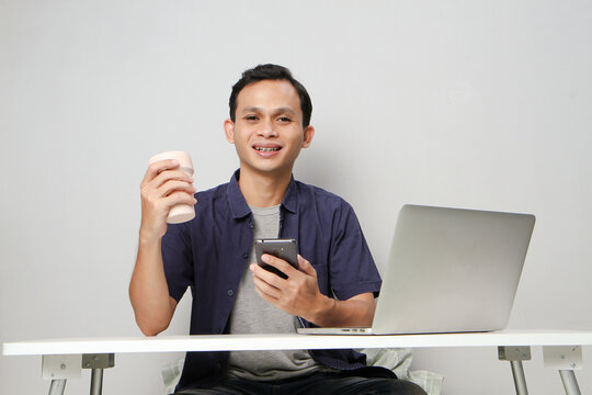 joyfull happy asian man holding mobile phone while sitting in front of laptop computer. on isolated background
