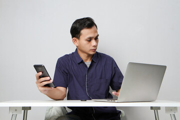 joyfull happy asian man holding mobile phone while sitting in front of laptop computer. on isolated background