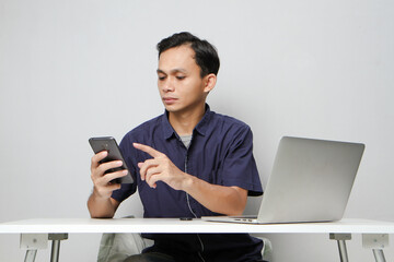 joyfull happy asian man holding mobile phone while sitting in front of laptop computer. on isolated background