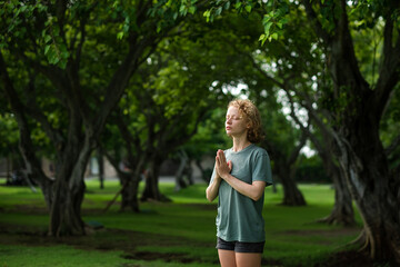 A young girl, with blond curly hair, folds her hands in namaste, meditates and thanks God, in the morning on a green meadow.
