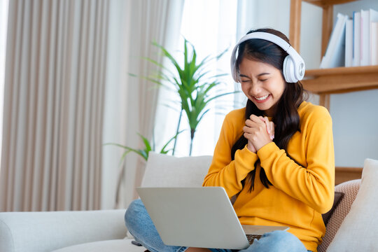 Young happy lucky woman student feeling excited winning, using computer laptop and sitting on sofa, adorable Asian female receiving great news on notebook, getting new job celebrating success
