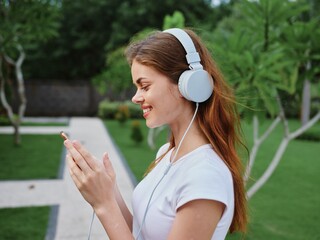 Woman wearing headphones listening to music through her phone smile with teeth walking in front of green palm trees on a summer trip