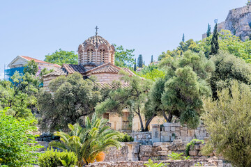 Lush greenery frames a Byzantine church set against a clear sky above historic ruins, in Athens, Greece