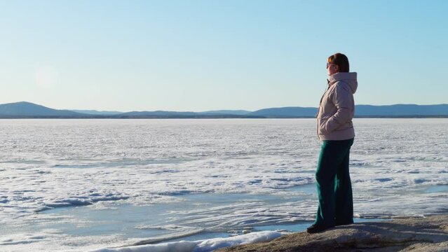 A woman with glasses stands alone on the shore of a frozen lake on a clear sunny day. A woman admires nature, getting real pleasure. Love of nature.