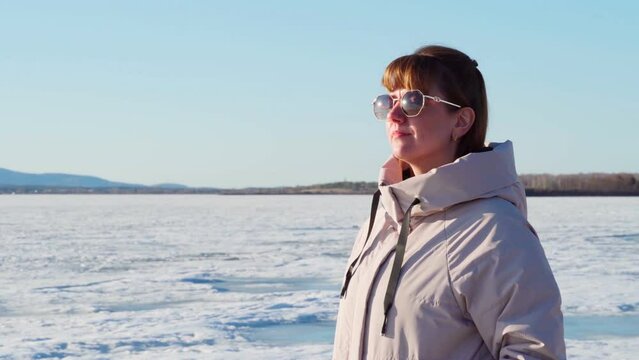 A woman with glasses stands alone on the shore of a frozen lake on a clear sunny day. A woman admires nature, getting real pleasure. Love of nature.