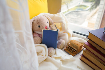 A toy hare is reading a book and sitting on the window in a white plaid
