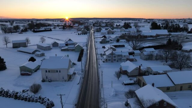 Snowy rural town at sunrise. American houses and farms under blanket of white snow during golden hour sunlight. Aerial rising shot.