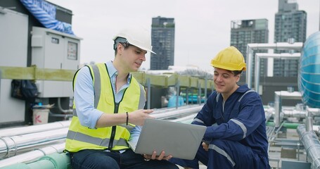 engineers manager and worker sitting on rooftop review plans on a laptop while on a construction site with high-rise buildings, indicating their different roles and responsibilities on the project
