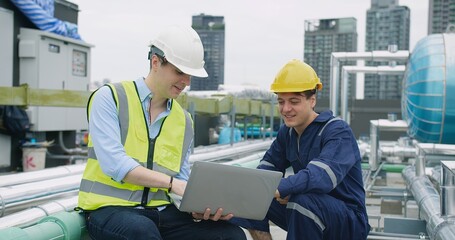 engineers manager and worker sitting on rooftop review plans on a laptop while on a construction site with high-rise buildings, indicating their different roles and responsibilities on the project