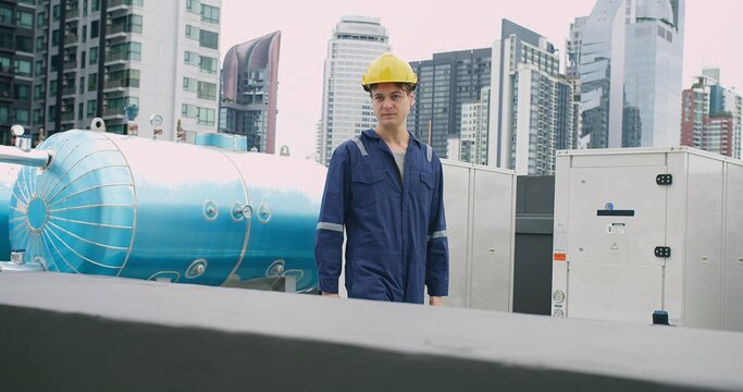 Engineer man in a blue jacket is walking inspecting maintenance insulated pipelines valve pump control on the roof at an industrial site near a large blue tank.