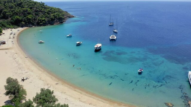 Aerial: Panoramic view of Tsougkria island beach with many tourist boats and sailboats. Tourists swim in turquoise clear blue water