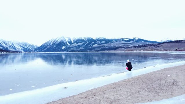 Photographer on the edge of a frozen lake with snow covered mountains in the background, aerial