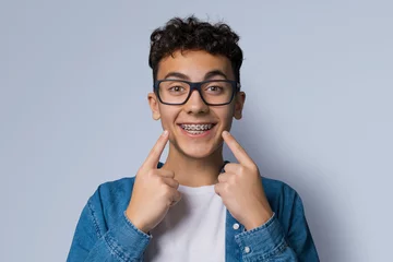  Dental dent care ad concept image - black сurly haired funny young man wear metal braces, eye glasses, show point white teeth smile. Isolated grey gray studio wall background. Positive optimistic. © vgstudio