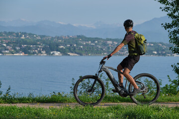 A man in a green T-shirt and black shorts with a backpack on a gray electric mountain bike in motion in the background Lake Garda, mountain peaks in the snow.