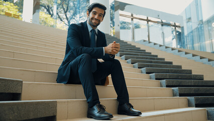 Smiling skilled businessman looking at camera while sitting on stairs. Young professional project...