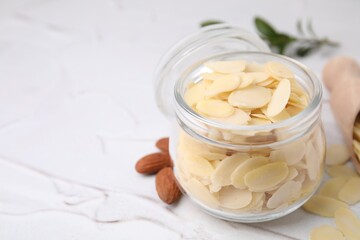 Fresh almond flakes in jar and nuts on white textured table, closeup. Space for text