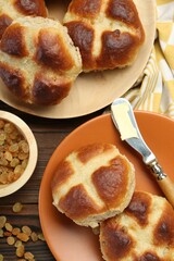 Tasty hot cross buns, knife with butter and raisins on wooden table, flat lay