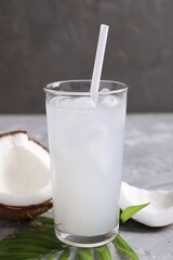 Glass of coconut water with ice cubes, palm leaf and nut on grey table