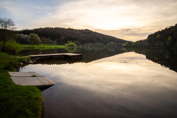A lake in a landscape shot. A sunset and the natural surroundings are reflected in the water of the reservoir. Marbachstausee, Odenwald, Hesse - obrazy, fototapety, plakaty