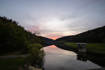 A lake in a landscape shot. A sunset and the natural surroundings are reflected in the water of the reservoir. Marbachstausee, Odenwald, Hesse - obrazy, fototapety, plakaty