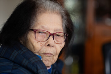 An elderly Asian woman turns to the camera and makes eye contact with a pensive expression. She is...
