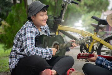 Asian boys hold guitar and playing near their bikes which parked by the narrow street in school area during their summer holiday, soft focus, summer activity of children concept.
